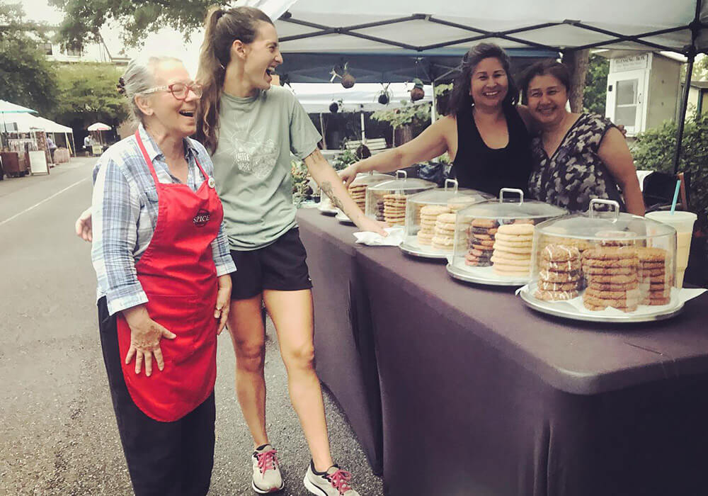 Bakers share a laugh behind a table of cookies under their market tent